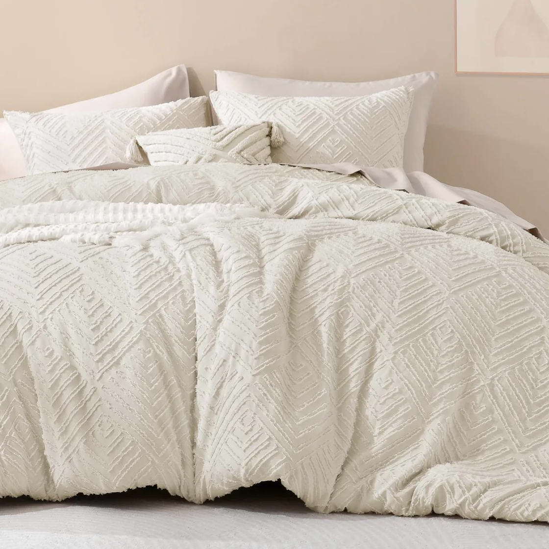 A textured off-white bedding set with zigzag patterns, including a duvet cover and pillows, on a bed in a neutral-toned room. A minimalist floral artwork hangs on the wall above.