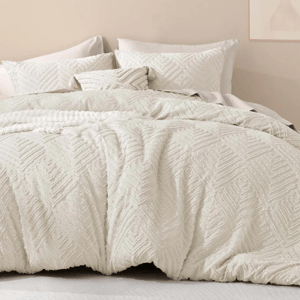 A textured off-white bedding set with zigzag patterns, including a duvet cover and pillows, on a bed in a neutral-toned room. A minimalist floral artwork hangs on the wall above.