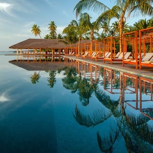A serene outdoor swimming pool surrounded by palm trees, with sun loungers under wooden canopies, offers a tranquil tropical setting. The clear blue sky and calm water create a reflective, peaceful ambiance.