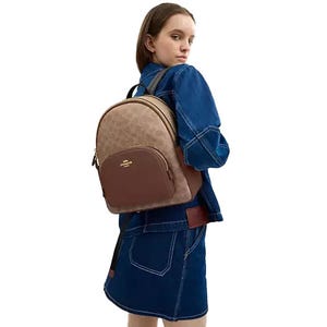 A person wearing a denim outfit is carrying a brown and beige patterned Coach backpack featuring the brand's logo and a front pocket.