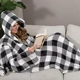 A person is cozily wrapped in a hooded black-and-white checkered blanket while reading a book on a couch.