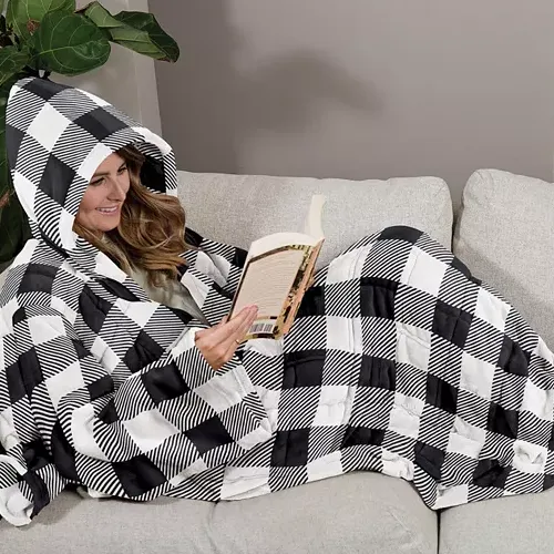 A person is cozily wrapped in a hooded black-and-white checkered blanket while reading a book on a couch.