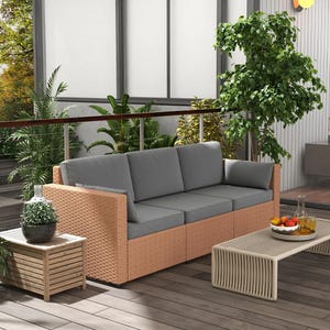 Outdoor patio scene featuring a wicker sofa with gray cushions, a wooden side table with a plant, a coffee table with fruit and a decanter, and lush green plants in the background.