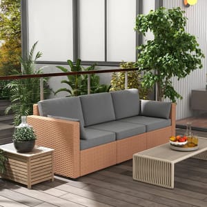 Outdoor patio scene featuring a wicker sofa with gray cushions, a wooden side table with a plant, a coffee table with fruit and a decanter, and lush green plants in the background.