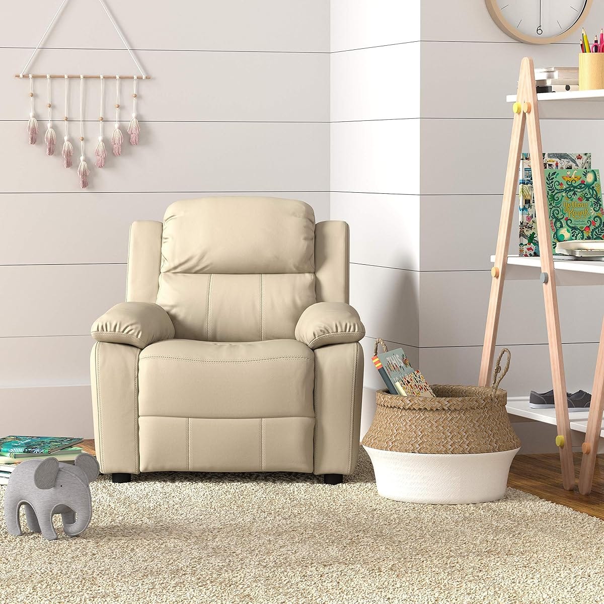 Beige recliner chair, white wall with hanging tassels, wooden easel with supplies, wicker basket, stuffed elephant, and a shaggy rug.