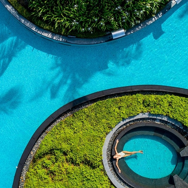 A person is lying on a curved sun deck by a swimming pool, surrounded by landscaped foliage.