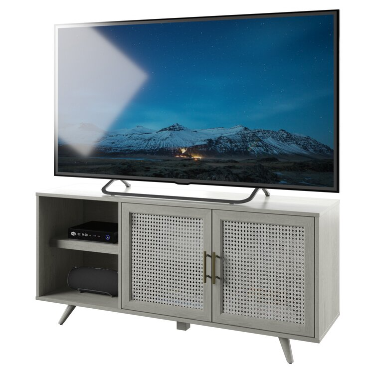 The Fairfax Oak TV Stand features a modern design with textured cabinet doors, open shelving for electronics, and angled legs. It's built to support a large flat-screen TV and offers storage and display space.