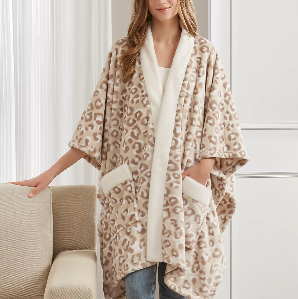 A person wearing a cozy, leopard-print fleece wrap with a white trim and pockets, paired with jeans, near a beige couch.