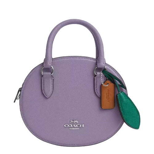 A lavender Coach handbag with a round shape, featuring dual handles and green and brown leather tags.