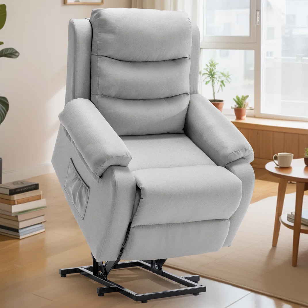 A gray lift recliner chair is shown in a living room setting, featuring padded armrests, a side pocket, and a mechanism for easy rise assistance.