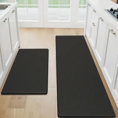 The photo shows two black kitchen mats on a wooden floor in a kitchen, positioned in front of the countertops and a stove area.