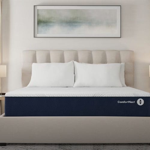A bed with a tufted headboard, featuring a ComfortNext branded mattress topped with white pillows, is set in a neutral-colored room with abstract wall art and a lamp on each side.