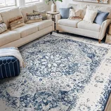 A living room features a beige sectional sofa with various textured cushions and a blue-and-white patterned area rug with a vintage design. A small side table and a navy pouf accent the space.