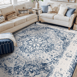 A living room features a beige sectional sofa with various textured cushions and a blue-and-white patterned area rug with a vintage design. A small side table and a navy pouf accent the space.