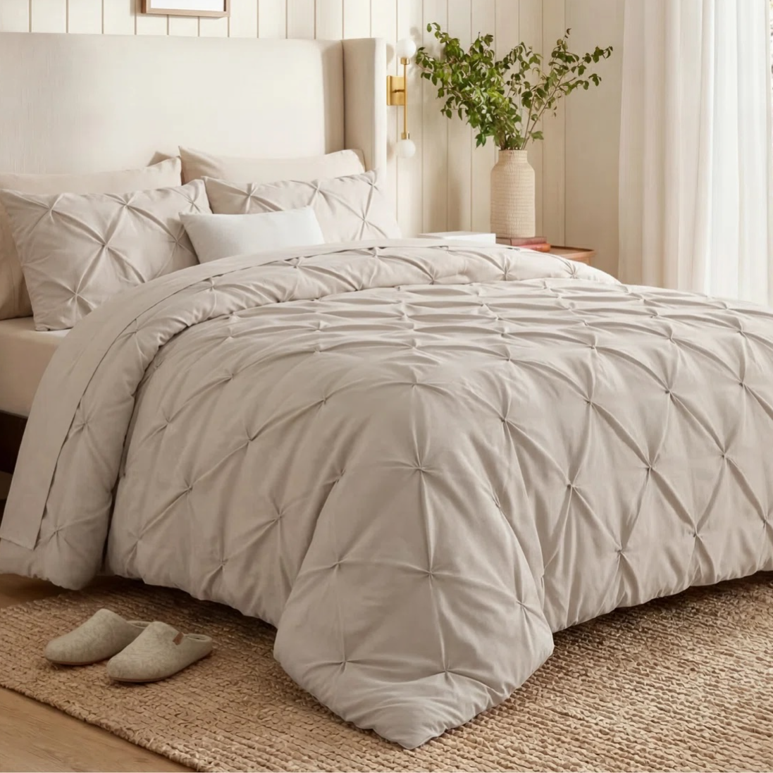 Cream-colored bedding set with a textured pin-tuck design on a bed, accompanied by a pair of slippers on a woven rug. Wall art and a potted plant add decor elements.