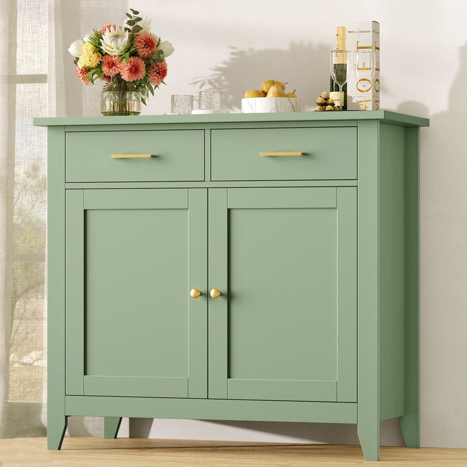 Green sideboard with two drawers and two cabinet doors, featuring gold handles and knobs. A vase of flowers, glasses, and a bowl of pears sit on top, along with a champagne bottle.