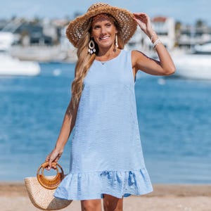 The Perfect Skies Mini Dress is a light blue, sleeveless dress featuring a textured fabric and a ruffled hem. It is styled with a woven straw hat and a straw handbag, conveying a relaxed, beach-ready look.