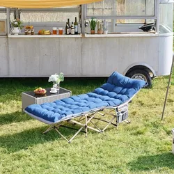 A blue folding camping recliner with a cushioned surface is placed on grass, next to a wicker side table with strawberries and a decorative plant. In the background, there's a food cart with various drinks and food items.