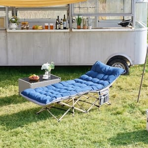 A blue folding camping recliner with a cushioned surface is placed on grass, next to a wicker side table with strawberries and a decorative plant. In the background, there's a food cart with various drinks and food items.