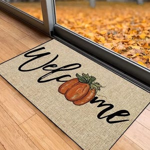 A welcome mat featuring a pumpkin design and the word \“Welcome\“ in cursive text, placed at a doorway with fallen leaves visible through the glass door.