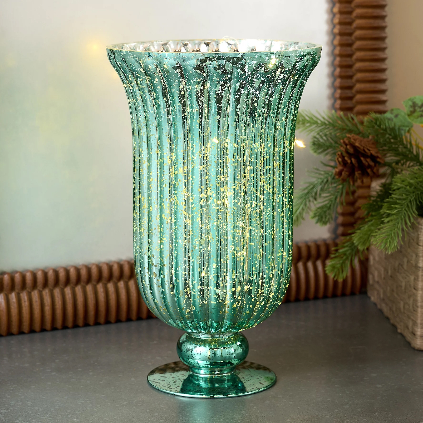 Tall, fluted teal glass hurricane vase with gold speckles and interior lighting. Decorated with a festive touch from surrounding pine branches and pinecones.