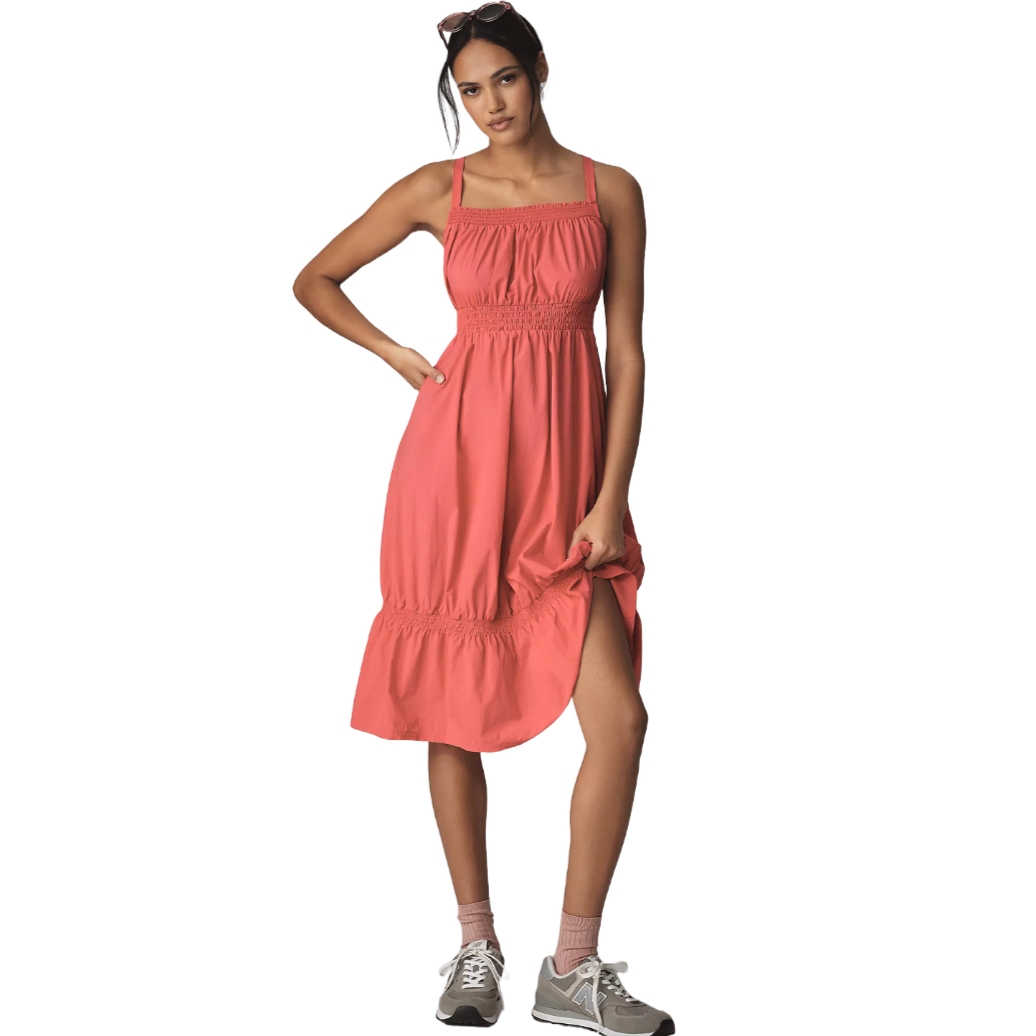 A woman is wearing a coral, sleeveless, ankle-length dress with a smocked waist and hem, paired with gray sneakers, pink socks, and sunglasses on her head.