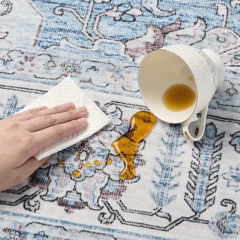 A hand uses a paper towel to clean up a spilled drink from a toppled teacup on a patterned rug.