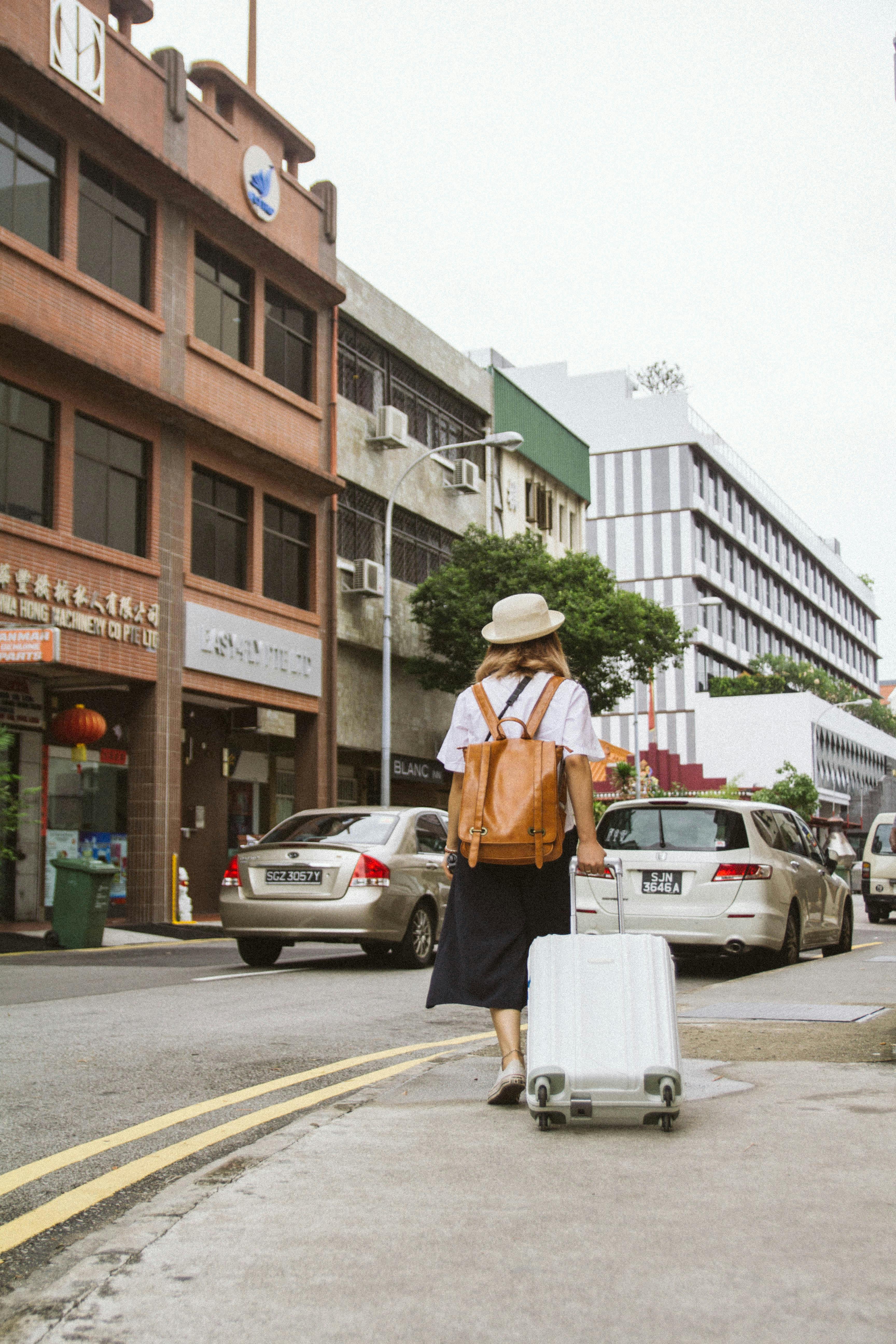 A person is walking along a city street holding a white suitcase and wearing a tan backpack.