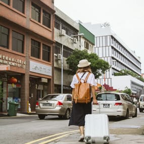 A person is walking along a city street holding a white suitcase and wearing a tan backpack.