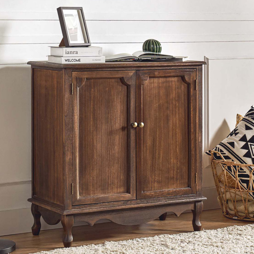 A wooden cabinet with brass handles is paired with a black floor lamp. There's a basket with geometric-patterned pillows nearby, and the cabinet surface holds books, a picture frame, and a small decorative item.