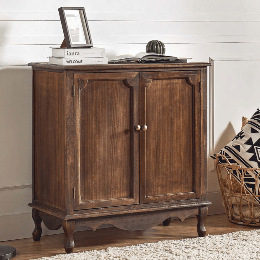 A wooden cabinet with brass handles is paired with a black floor lamp. There's a basket with geometric-patterned pillows nearby, and the cabinet surface holds books, a picture frame, and a small decorative item.