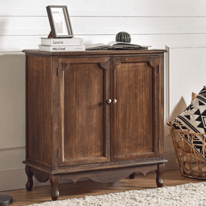 A wooden cabinet with brass handles is paired with a black floor lamp. There's a basket with geometric-patterned pillows nearby, and the cabinet surface holds books, a picture frame, and a small decorative item.