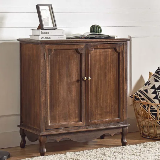 A wooden cabinet with brass handles is paired with a black floor lamp. There's a basket with geometric-patterned pillows nearby, and the cabinet surface holds books, a picture frame, and a small decorative item.