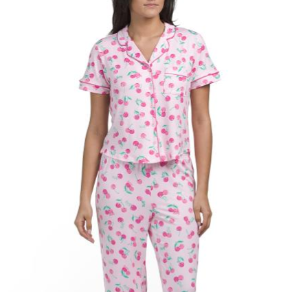 A woman is wearing pink pajamas with a cherry pattern, featuring a short-sleeved button-up top and long pants.