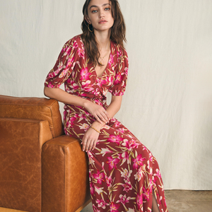 A woman is seated wearing a deep v-neck floral dress with hoop earrings and a gold bracelet.