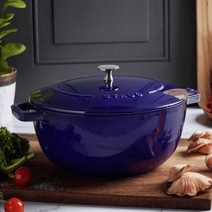 A blue Staub cast iron cocotte with a lid and a metal knob, surrounded by vegetables and mushrooms on a wooden board.