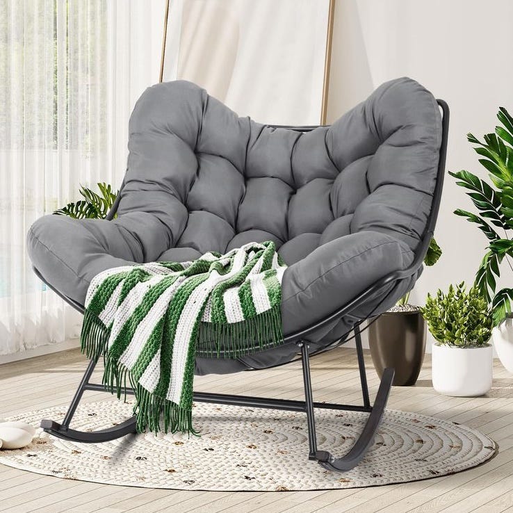 A gray padded rocking chair with a plush, tufted design and a metal frame, adorned with a green and white striped blanket, is placed on a circular woven rug surrounded by potted plants.