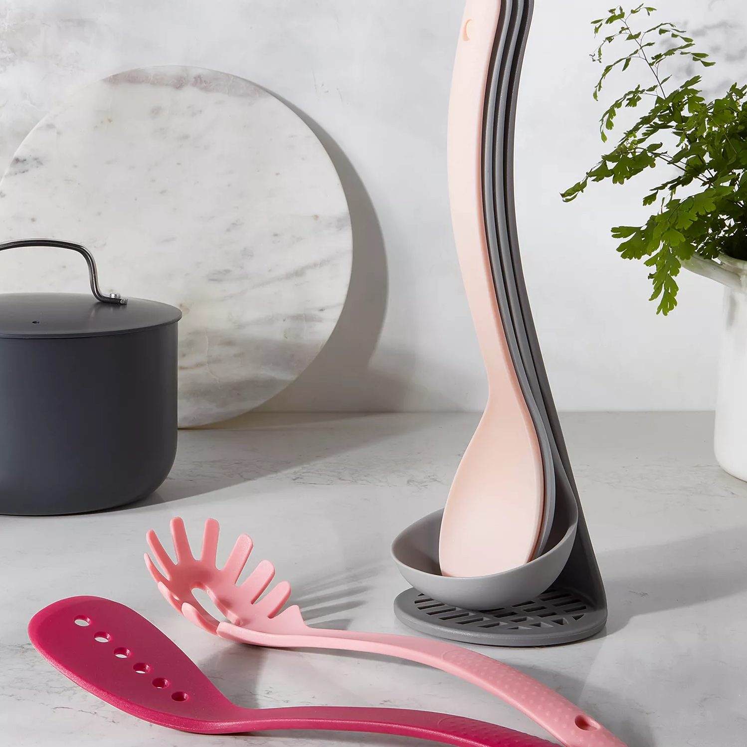 A set of nested pink and gray kitchen utensils on a countertop, with a cooking pot and a potted plant in the background.