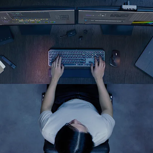 A person is working at a desk with two monitors displaying editing software. On the desk are a keyboard, mouse, external hard drive, and some small electronic accessories.