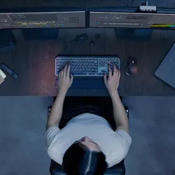 A person is working at a desk with two monitors displaying editing software. On the desk are a keyboard, mouse, external hard drive, and some small electronic accessories.