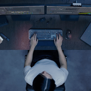 A person is working at a desk with two monitors displaying editing software. On the desk are a keyboard, mouse, external hard drive, and some small electronic accessories.