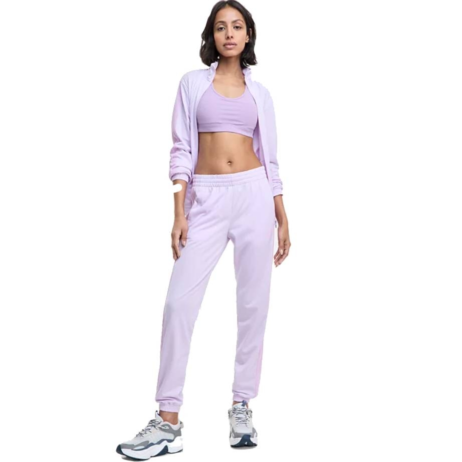 Light purple Adidas Tricot 3-Stripes Track Pants paired with a matching jacket and sports bra, featuring a relaxed fit with elastic waistband and cuffs. Accompanied by white and black athletic sneakers.