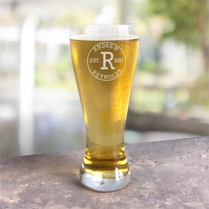 A clear pilsner glass filled with beer features personalized engraved text, including \“Andrew Reynolds,\“ \“EST. 2003,\“ and a large \“R\“ in the center, all within a circular emblem design.