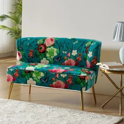 A small loveseat with a vibrant floral pattern on a teal background, featuring gold legs. The setting includes a round side table with a lamp and an open book, on a light-colored floor with a rug.