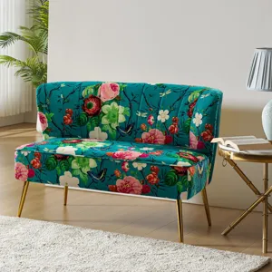 A small loveseat with a vibrant floral pattern on a teal background, featuring gold legs. The setting includes a round side table with a lamp and an open book, on a light-colored floor with a rug.