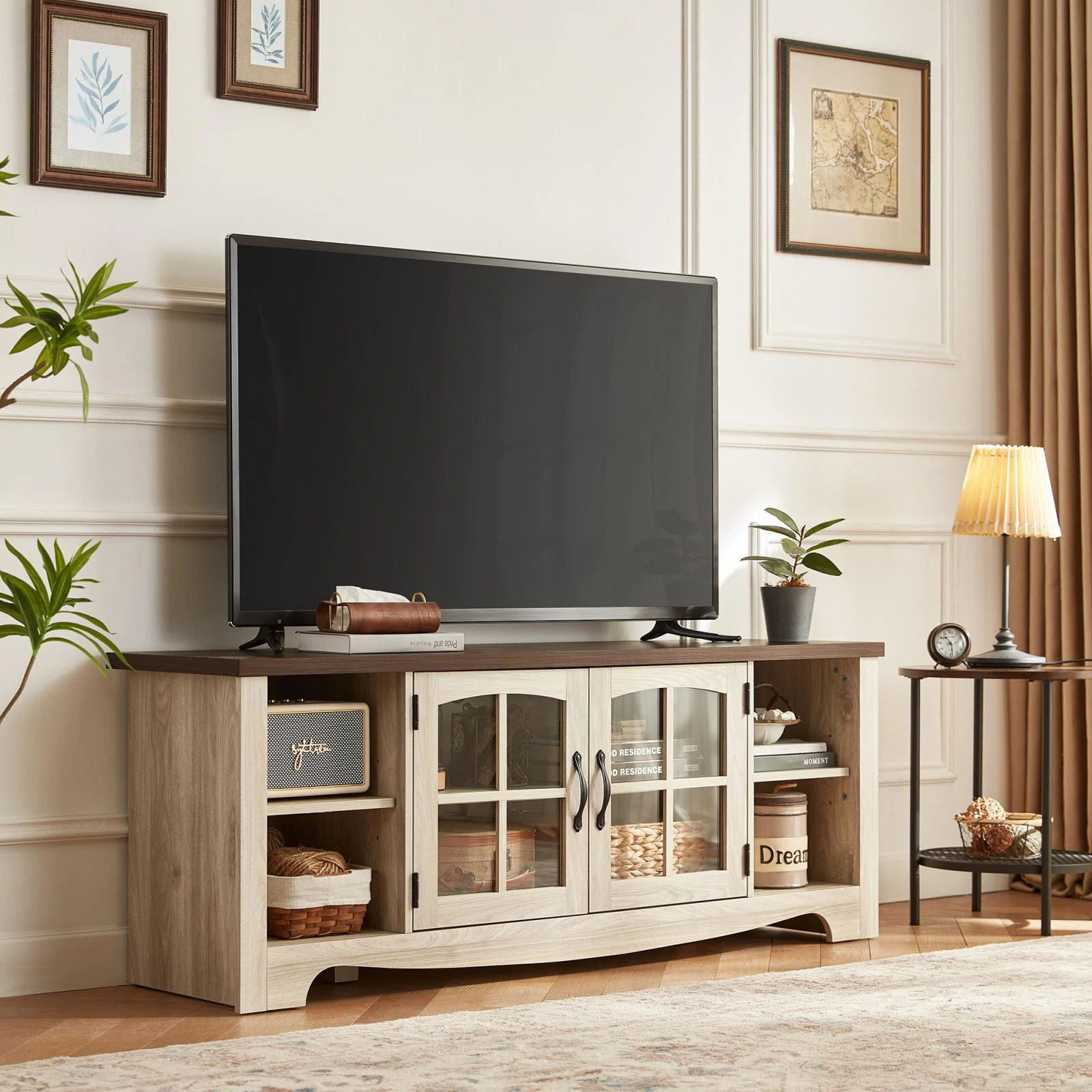 A flat-screen TV sits on a wooden TV stand with glass doors and decorative items, in a room with plants and framed art.