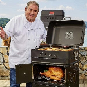 A man in a chef's coat stands beside an open outdoor grill with a roasted chicken inside.