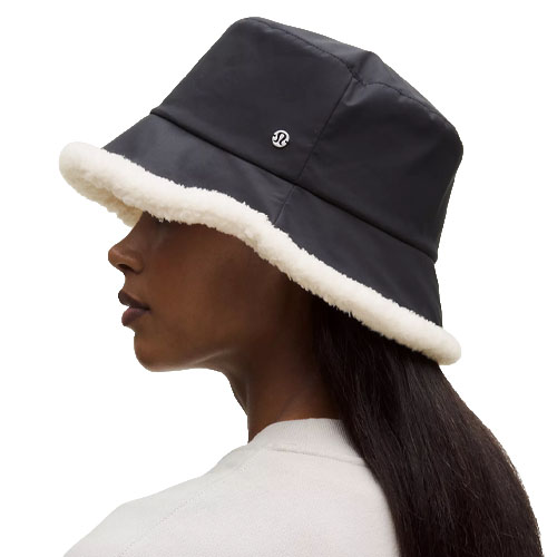 A black bucket hat with a white faux fur trim and a small logo on the side, worn by a person with long dark hair.