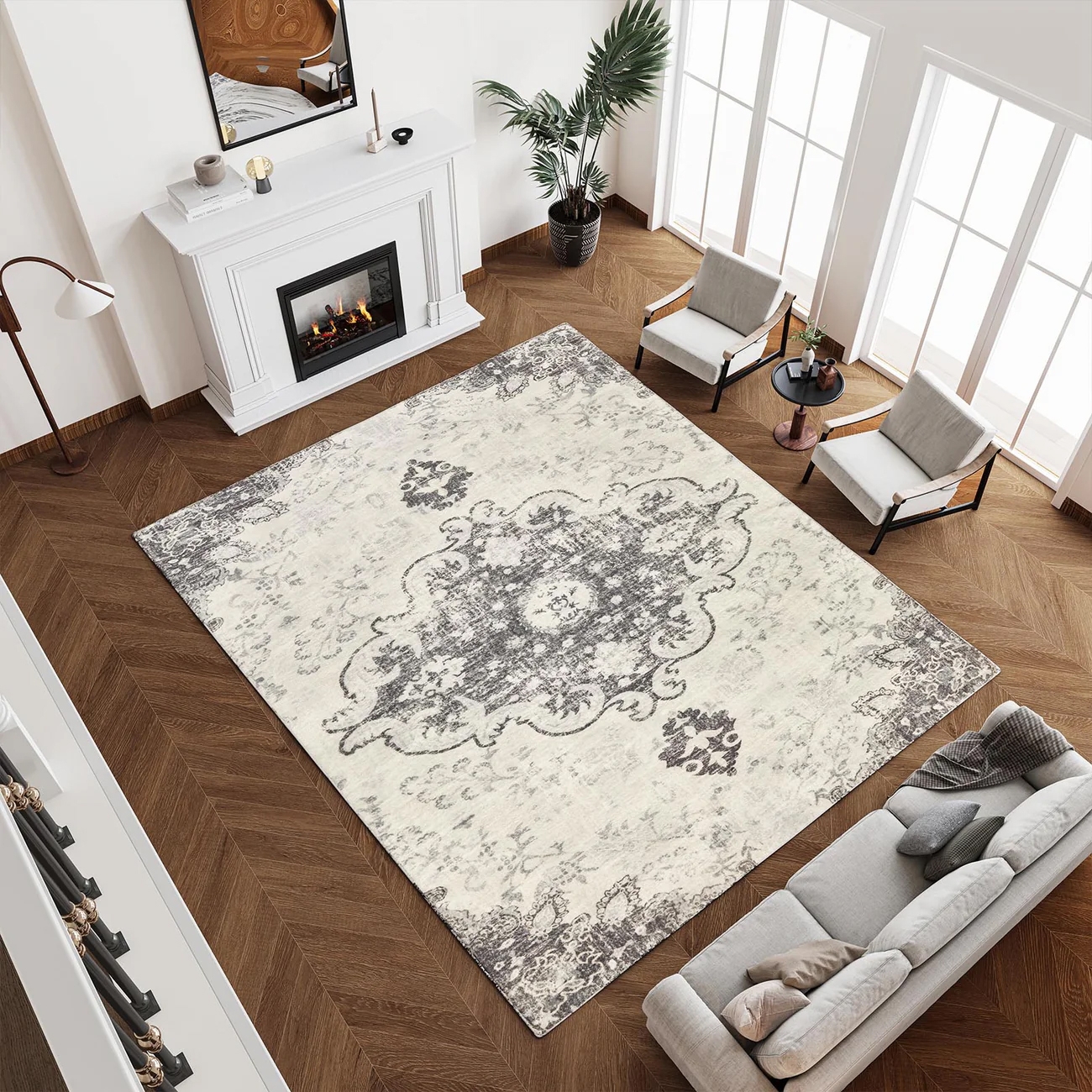 A living room with a large, patterned area rug on a herringbone wood floor, a gray sofa, two armchairs with a small round table, a white fireplace with a mirror above, and a tall floor lamp.