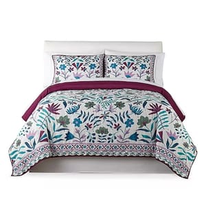A bed set featuring a quilt and two shams with a colorful floral pattern in shades of teal, purple, and green on a white background, accompanied by a deep purple inner lining.